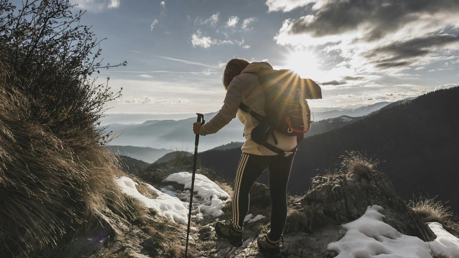 A hiker with a backpack and walking stick stands on a rocky, snow-dusted mountain slope at sunset, overlooking distant peaks and hiking trails in the Smoky Mountains under a partly cloudy sky.
