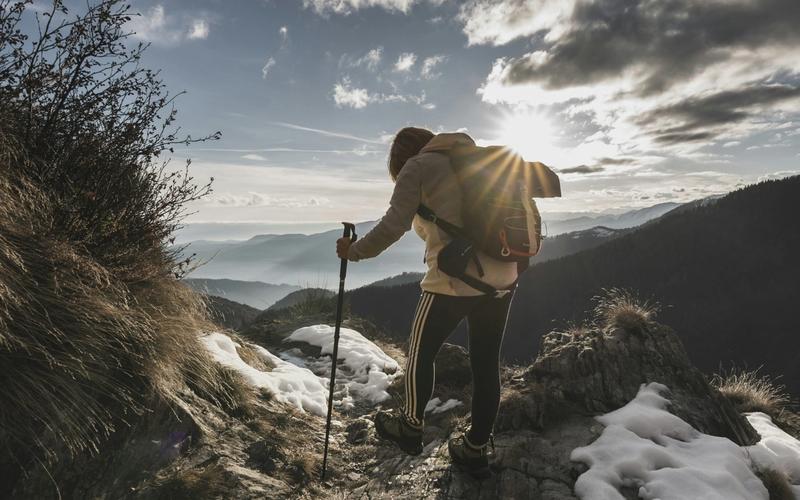 A hiker with a backpack and walking stick stands on a rocky, snow-dusted mountain slope at sunset, overlooking distant peaks and hiking trails in the Smoky Mountains under a partly cloudy sky.