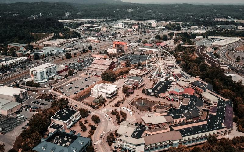 Aerial view of Pigeon Forge, showcasing its iconic Ferris wheel at the center, surrounded by city attractions, buildings, parking lots, roads, and distant green hills under a hazy sky.
