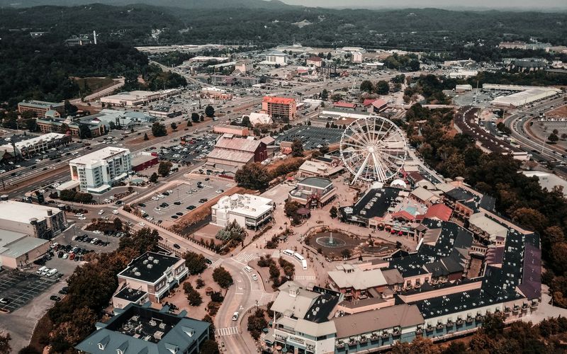 Aerial view of Pigeon Forge, showcasing its iconic Ferris wheel at the center, surrounded by city attractions, buildings, parking lots, roads, and distant green hills under a hazy sky.