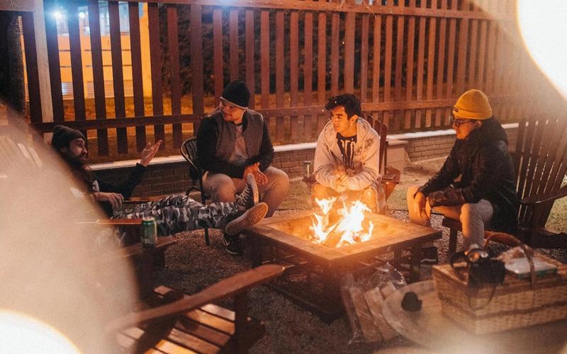Four people sit around a fire pit outdoors at night, relaxing on wooden chairs after a day exploring the Smoky Mountains. Dressed warmly in jackets and hats, they chat casually by the fence, enjoying their cozy corporate retreat.
