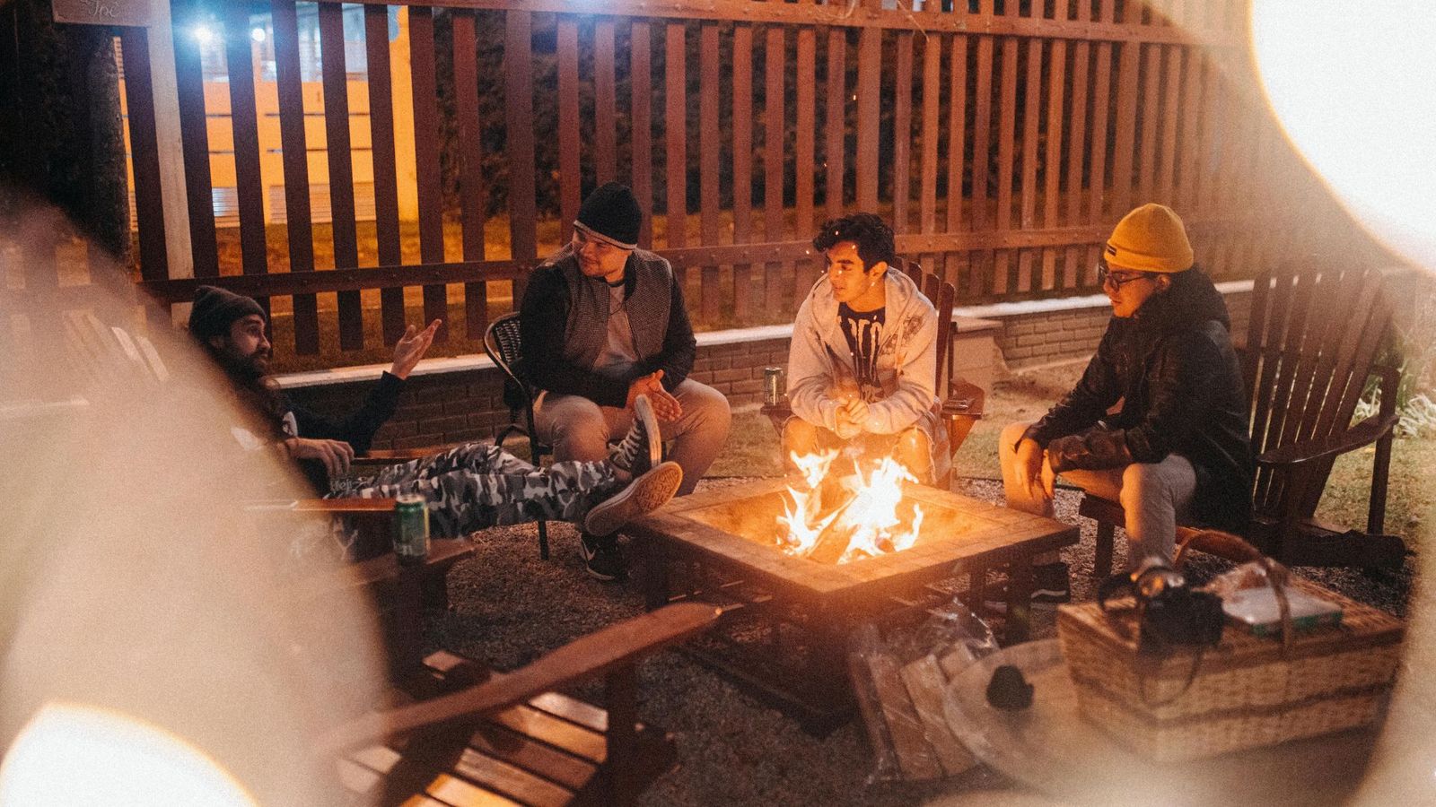 Four people sit around a fire pit outdoors at night, relaxing on wooden chairs after a day exploring the Smoky Mountains. Dressed warmly in jackets and hats, they chat casually by the fence, enjoying their cozy corporate retreat.