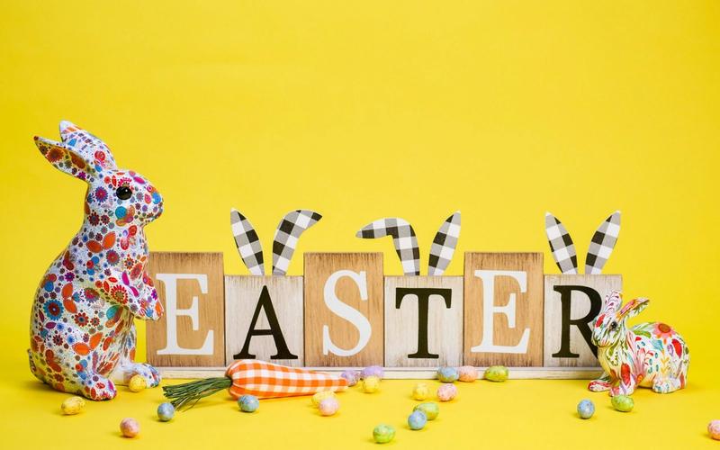 Colorful decorative rabbits and wooden blocks spelling EASTER with bunny ear decorations sit on a bright yellow background, surrounded by pastel candies and two fabric carrots—perfect inspiration for Smoky Mountains Easter activities.