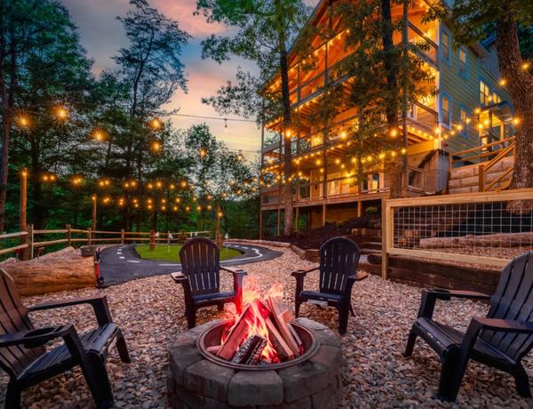 A cozy outdoor fire pit is surrounded by Adirondack chairs, string lights, and trees, with a multi-story cabin in the background at sunset, creating a warm, inviting atmosphere.