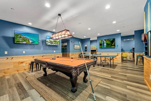 A cozy game room in a Smoky Mountain cabin features a pool table, ping pong table, photos on blue walls, two TVs, and wood panel accents. The wood floor is complemented by a rug under the pool table, while ceiling lights illuminate this inviting space—perfect for gathering with friends and communities.