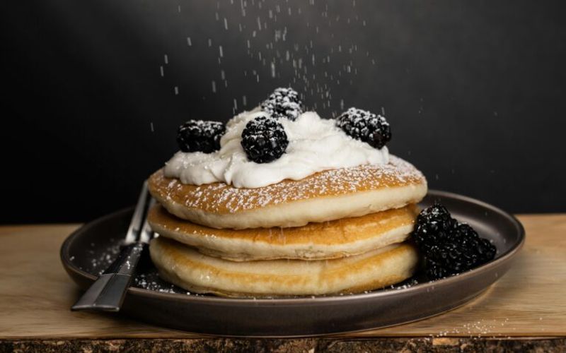 A stack of pancakes topped with whipped cream and blackberries, being sprinkled with powdered sugar, sits on a dark plate—just like those you’ll find on the Log Cabin Pancake House menu in Gatlinburg TN. A fork rests beside it against a dark background.