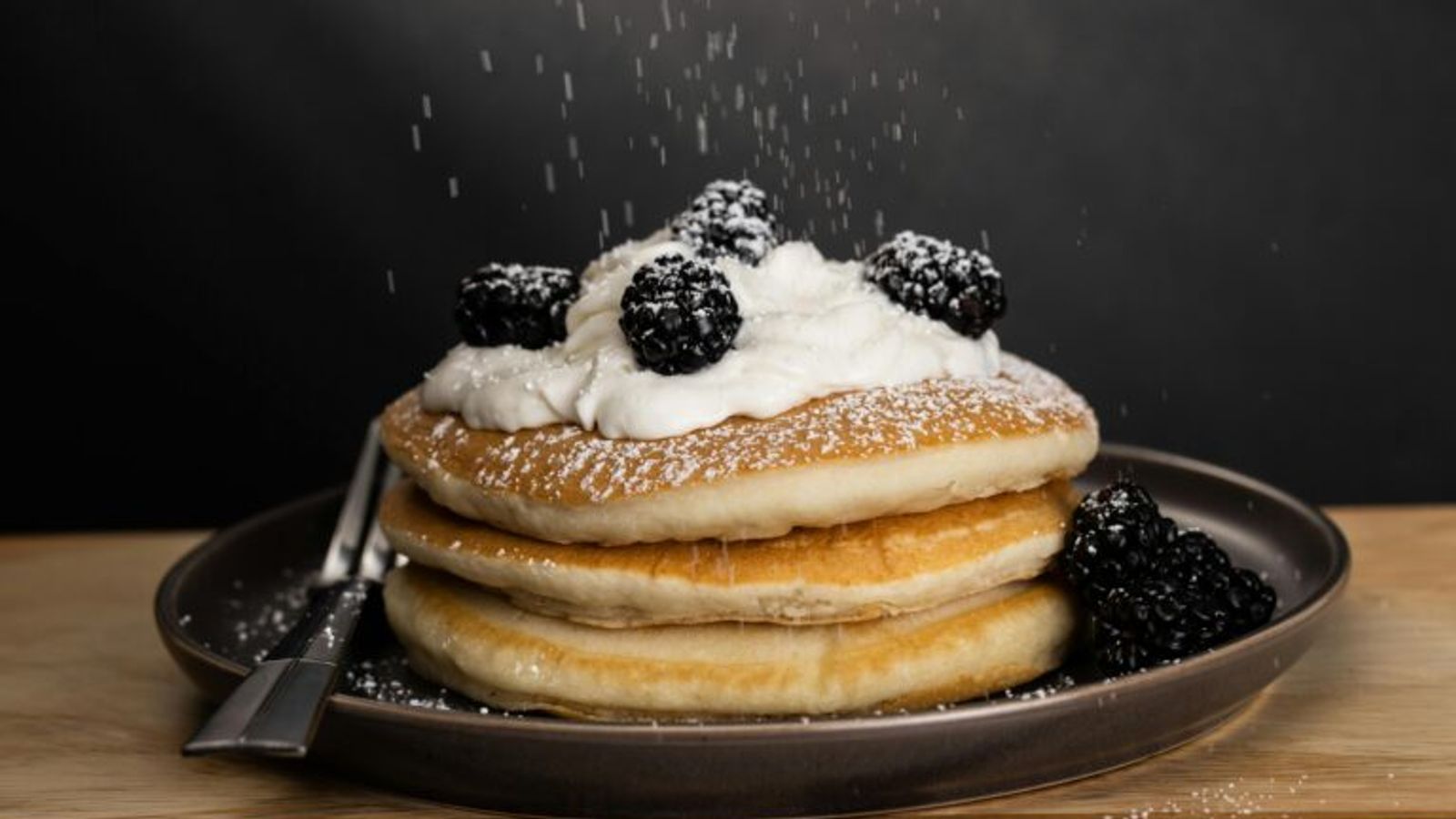 A stack of pancakes topped with whipped cream and blackberries, being sprinkled with powdered sugar, sits on a dark plate—just like those you’ll find on the Log Cabin Pancake House menu in Gatlinburg TN. A fork rests beside it against a dark background.