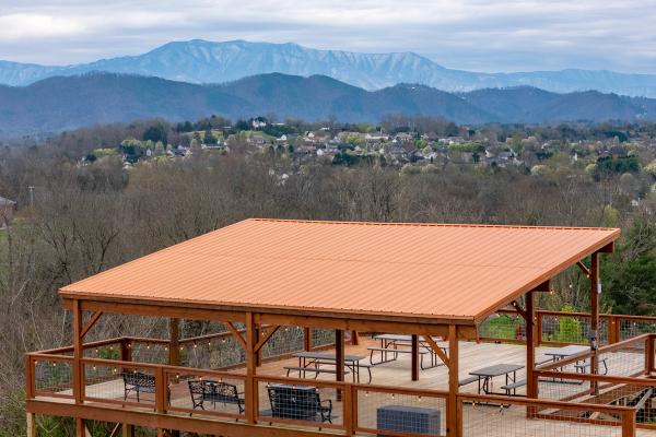 Wooden pavilion with orange metal roof, picnic tables, and string lights overlooks a valley of Smoky Mountain cabin communities, dotted with houses and trees against distant mountains under a cloudy sky.