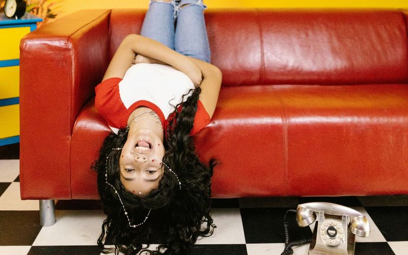 A person with long dark hair lies upside down on a red couch at WonderWorks Pigeon Forge, wearing a red and white shirt and blue jeans. The floor is black-and-white checkered, with an old-fashioned rotary phone nearby. Check hours and tickets before you visit!.
