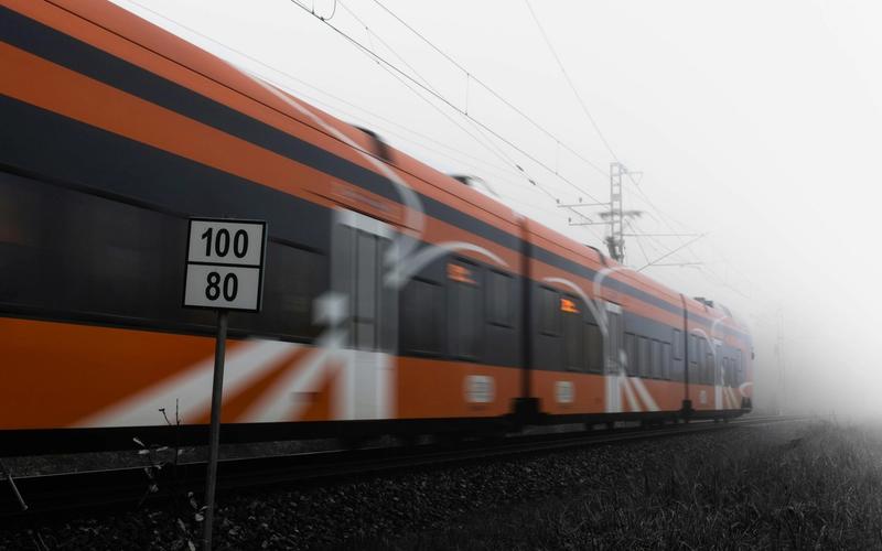 An orange train speeds past on railway tracks in a foggy setting, with a sign in the foreground displaying speed limits of 100 and 80—perfect inspiration for travel tips or things to do while exploring Gatlinburg in winter.