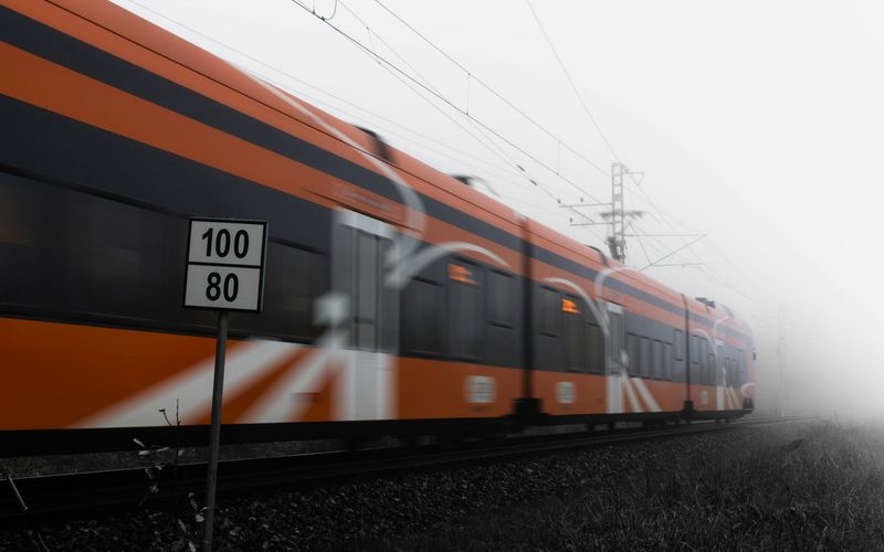An orange train speeds past on railway tracks in a foggy setting, with a sign in the foreground displaying speed limits of 100 and 80—perfect inspiration for travel tips or things to do while exploring Gatlinburg in winter.
