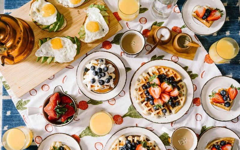 A breakfast table inspired by the best breakfast places in Gatlinburg, set with waffles topped with berries, avocado toast with fried eggs, orange juice, coffee, and a bowl of strawberries on a strawberry-patterned tablecloth.