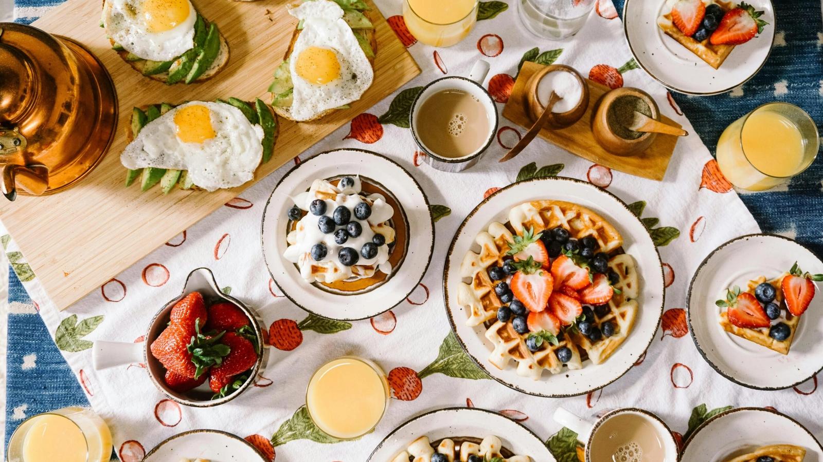 A breakfast table inspired by the best breakfast places in Gatlinburg, set with waffles topped with berries, avocado toast with fried eggs, orange juice, coffee, and a bowl of strawberries on a strawberry-patterned tablecloth.