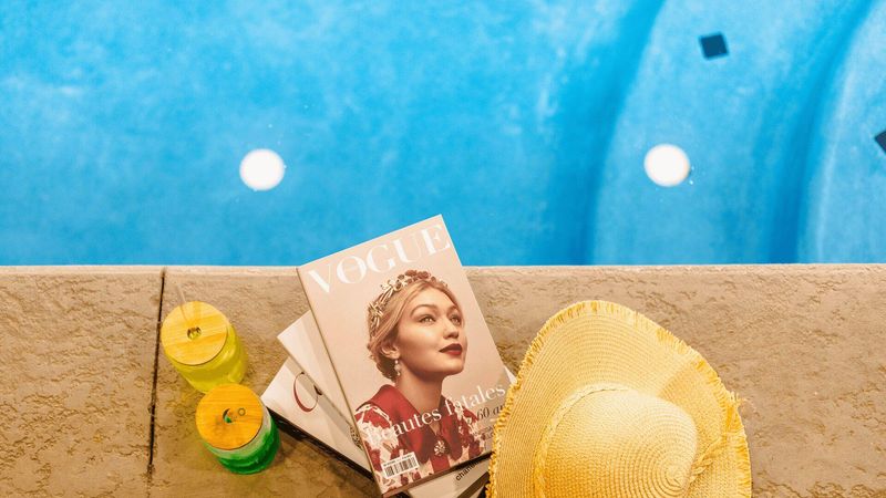 A Vogue magazine next to a pool, with a sun hat and two drinks.
