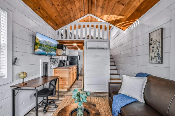 A bright tiny house interior with wood accents evokes the charm of Smoky Mountain cabin communities. Featuring a loft above, a compact kitchen, a wall-mounted TV, a small desk with a globe, a brown sofa with a blue throw, and a round coffee table with a vase, this inviting space captures the warmth and spirit of the Smoky Mountain lifestyle.