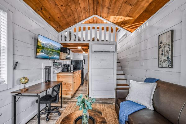 A bright tiny house interior with wood accents evokes the charm of Smoky Mountain cabin communities. Featuring a loft above, a compact kitchen, a wall-mounted TV, a small desk with a globe, a brown sofa with a blue throw, and a round coffee table with a vase, this inviting space captures the warmth and spirit of the Smoky Mountain lifestyle.