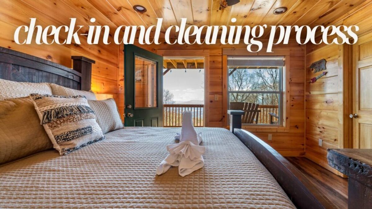 A cozy wooden cabin bedroom with a neatly made bed, decorative pillows, and a towel folded like a swan. Large windows reveal trees and a porch outside. Text reads, check-in and cleaning process—discover Why We Stand Out.