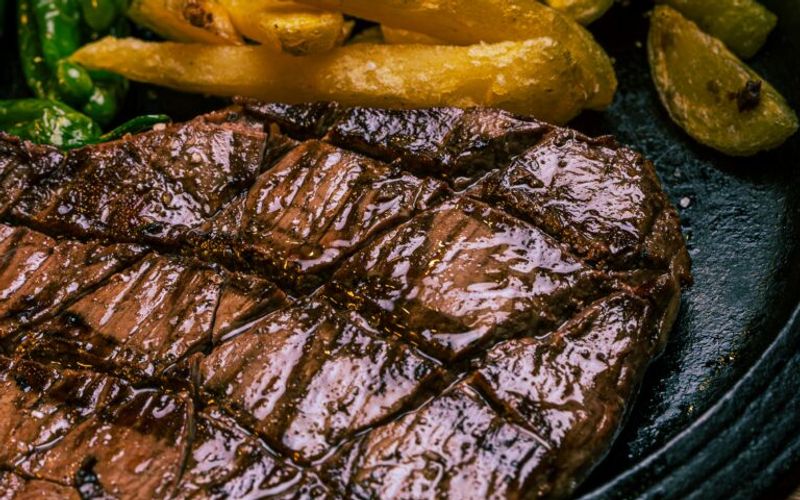 Close-up of a grilled steak with grill marks, served with golden French fries and green vegetables on a dark plate—just like you'd find on the Peddler Steakhouse menu. The steak appears juicy and cooked to perfection.