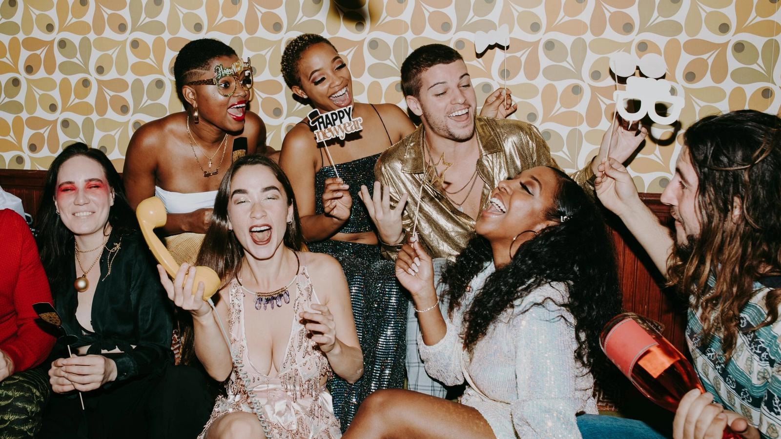 A group of friends dressed in festive outfits laugh and celebrate New Year's Eve together indoors in Gatlinburg, TN, holding party props and noisemakers against a retro-patterned wallpaper background.