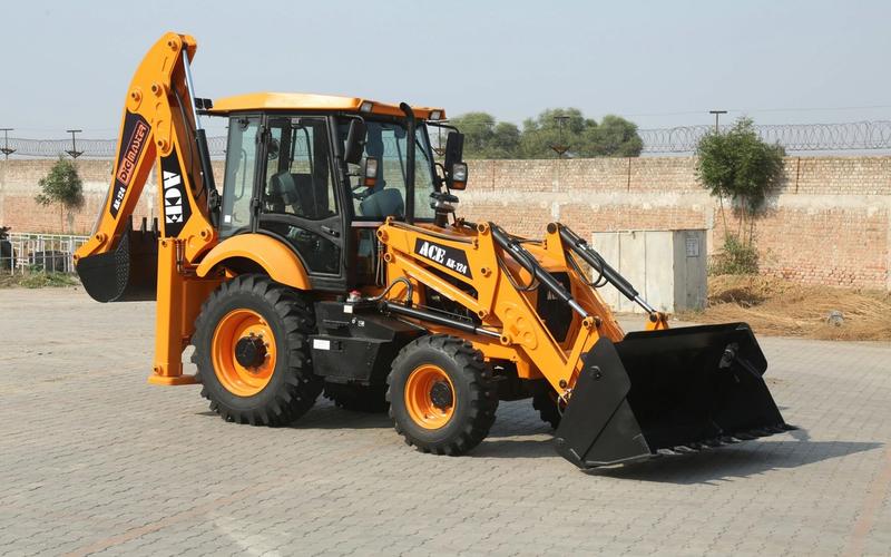 A yellow ACE backhoe loader is parked on a paved surface, featuring a front bucket and rear digging arm—just like the equipment you’ll see at Dig’n Zone Theme Park—with a brick wall and trees in the background.