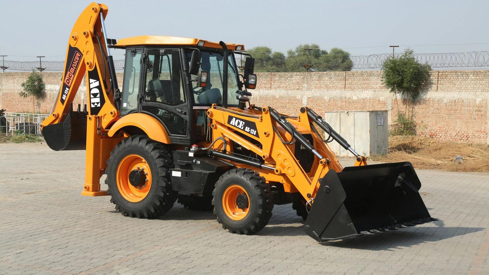 A yellow ACE backhoe loader is parked on a paved surface, featuring a front bucket and rear digging arm—just like the equipment you’ll see at Dig’n Zone Theme Park—with a brick wall and trees in the background.