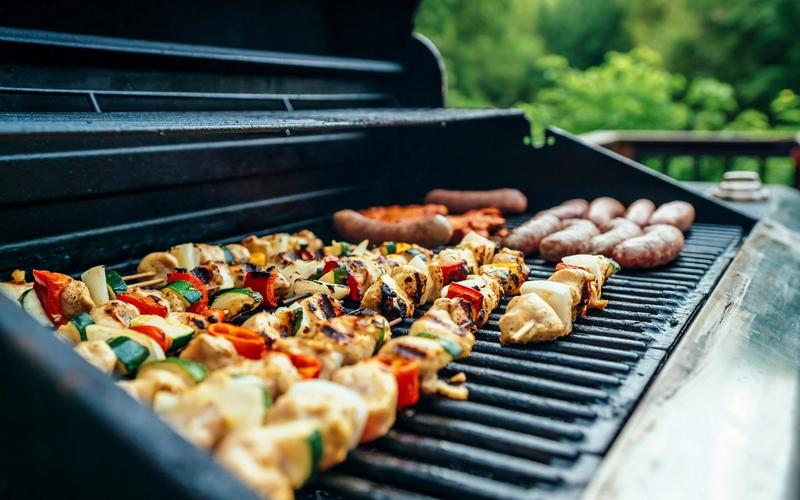Colorful vegetable and chicken skewers, as well as sausages, sizzle on a Smokies BBQ, grilling outdoors with green trees visible in the blurred background.