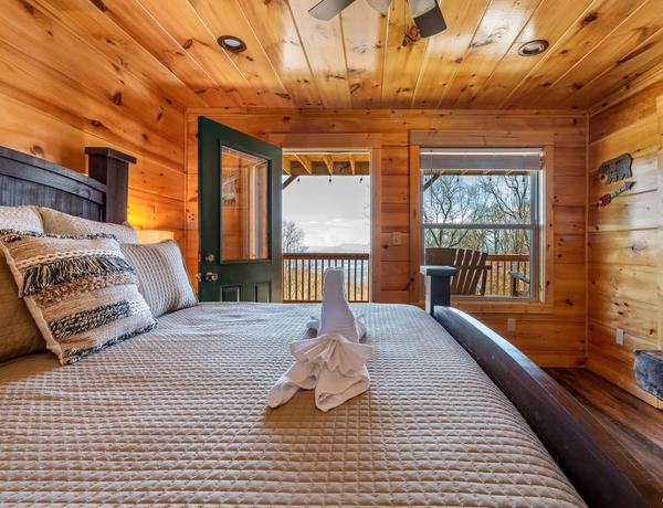 A cozy wooden cabin bedroom with a neatly made bed, folded towel art, open door, and large window showcasing a scenic view of trees and mountains outside.