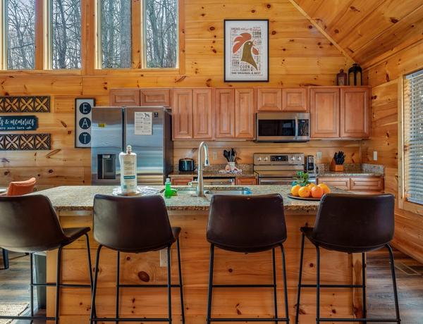 A cozy, modern kitchen with wood-paneled walls, stainless steel appliances, a large island with four dark chairs, fruit and soap on the counter, and a large window letting in sunlight.