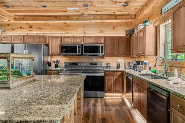 Modern kitchen with wood-paneled walls reminiscent of cozy Smoky Mountain cabins, stainless steel appliances, granite countertops, a double oven, a large sink under a window, and various kitchen gadgets and utensils neatly arranged on the counters.