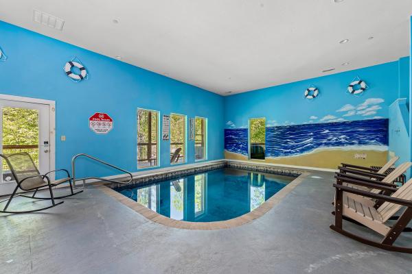 Indoor pool room in a Smoky Mountain Cabin, featuring a blue ocean-themed mural, life preservers on the wall, safety sign, several windows, a door leading outside, and rocking chairs arranged around the pool.
