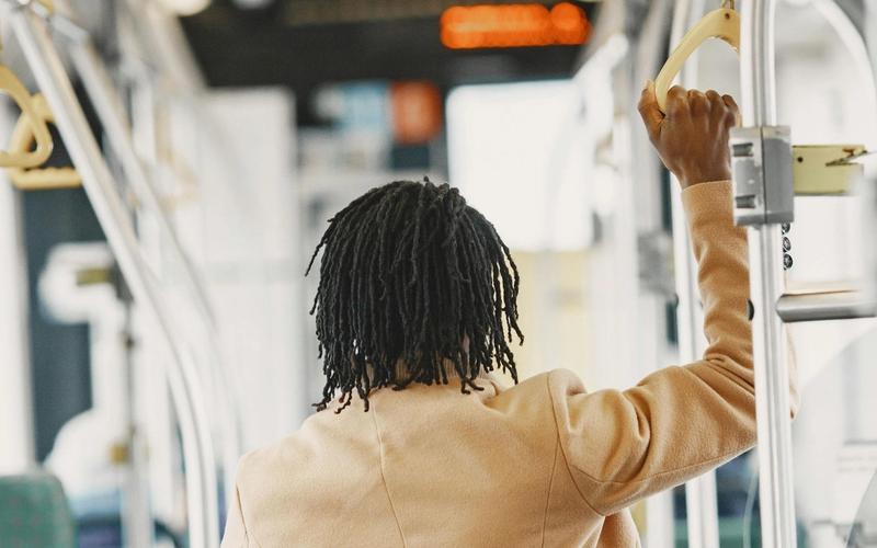 A person with short, twisted hair wearing a tan coat stands inside a Gatlinburg trolley, holding onto a metal handrail. The individual is facing away from the camera, experiencing convenient Gatlinburg transportation.