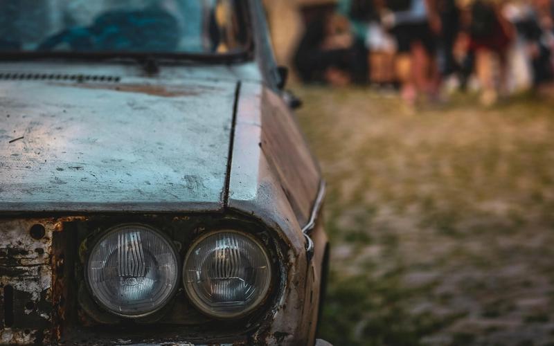 Close-up of the front left side of an old, rusty car with two round headlights, parked on a cobblestone road. The blurred background hints at people mingling, capturing the authentic vibe often found at Pigeon Forge car shows.