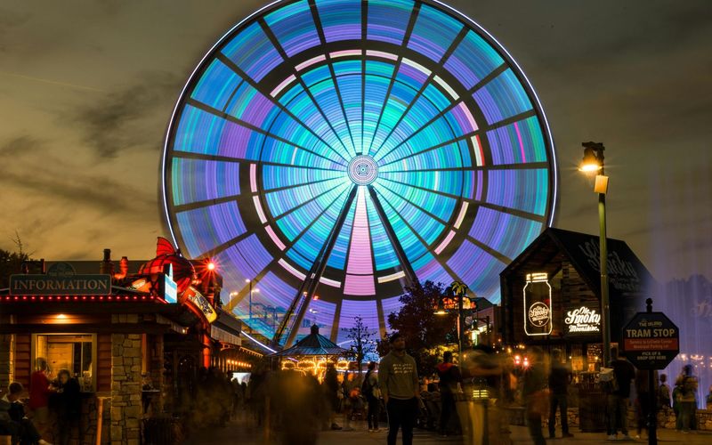 A large Ferris wheel with colorful, glowing lights spins above The Island in Pigeon Forge at dusk, where people stroll past illuminated signs and a nearby tram stop—one of the must-see Pigeon Forge attractions.