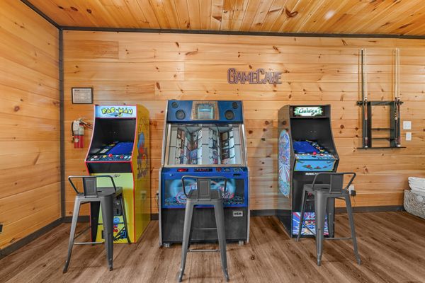 Three arcade machines—Pac-Man, a racing game, and Galaga—are lined up against a wooden wall in a cozy cabin game room. Three metal stools are placed in front of the machines, and pool cues hang on the right, creating the perfect spot for fun and relaxation in the heart of Smoky Mountain communities.