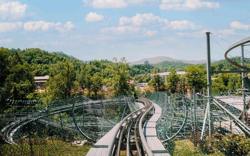 A view from the front seat of the Smoky Mountain Alpine Coaster track winding through green trees and hills, with blue sky and scattered clouds in the background.