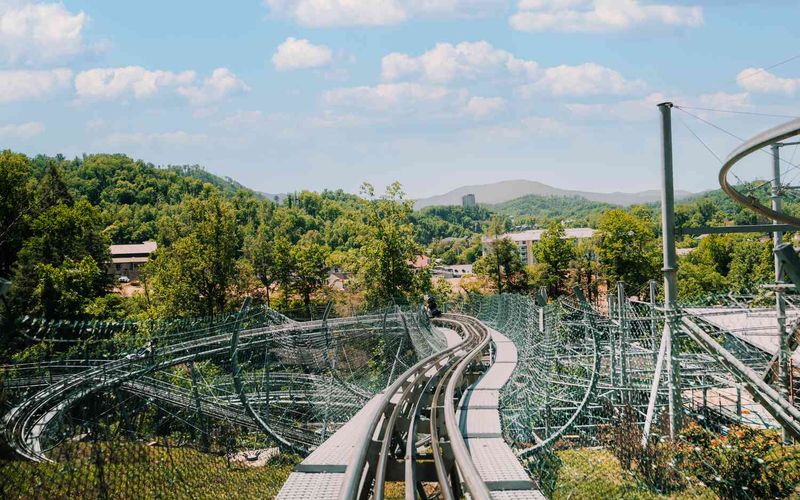 A view from the front seat of the Smoky Mountain Alpine Coaster track winding through green trees and hills, with blue sky and scattered clouds in the background.