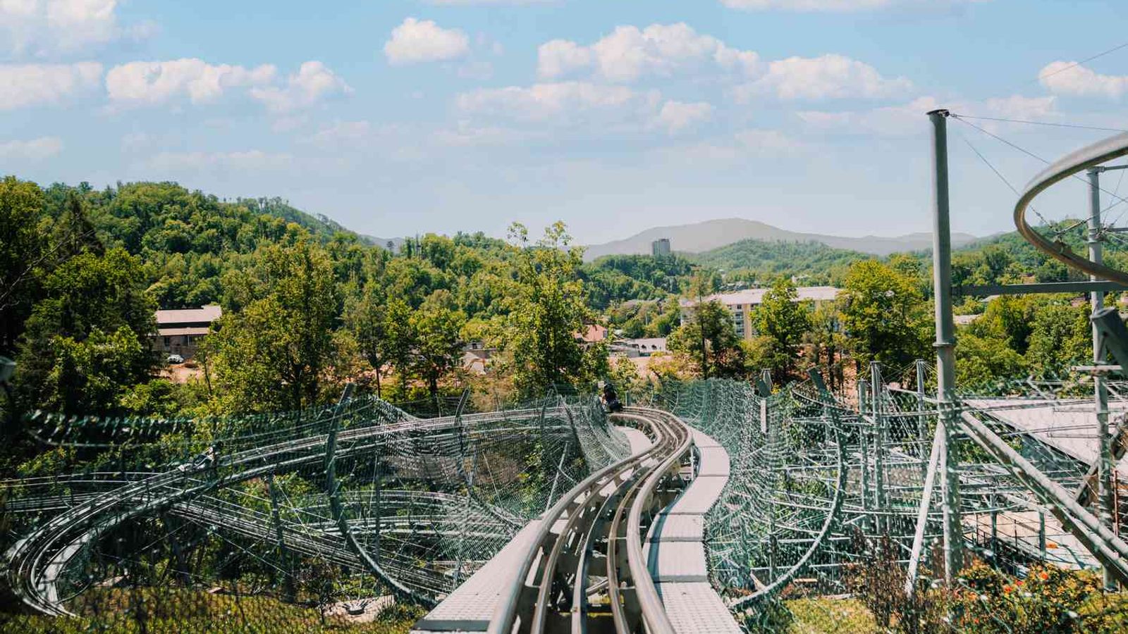 A view from the front seat of the Smoky Mountain Alpine Coaster track winding through green trees and hills, with blue sky and scattered clouds in the background.