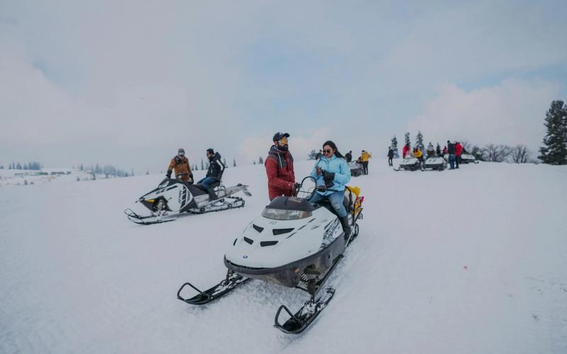 People enjoying winter activities on snowmobiles in a snowy landscape near Gatlinburg, TN. Dressed in winter clothing, some riders are moving while others are stationary among trees under a cloudy sky.