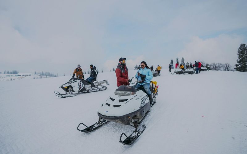 People enjoying winter activities on snowmobiles in a snowy landscape near Gatlinburg, TN. Dressed in winter clothing, some riders are moving while others are stationary among trees under a cloudy sky.