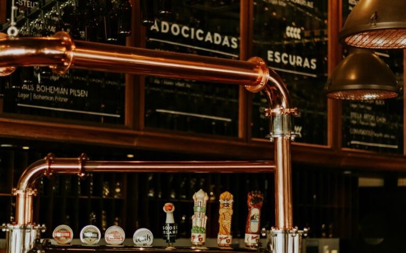 Beer taps with various labels are displayed on a polished copper bar in Gatlinburg. Glasses hang above, and a chalkboard menu with categories in Portuguese is visible in the background under warm lighting, evoking local distilleries.