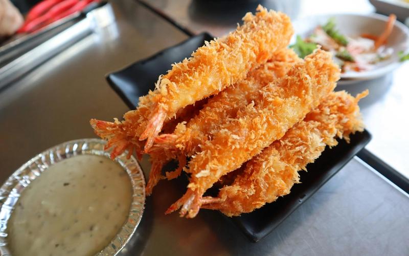A black plate holds several pieces of golden, crispy fried shrimp from the Bubba Gump menu, with a foil dish of creamy dipping sauce nearby. In the background, there is a small plate of salad.