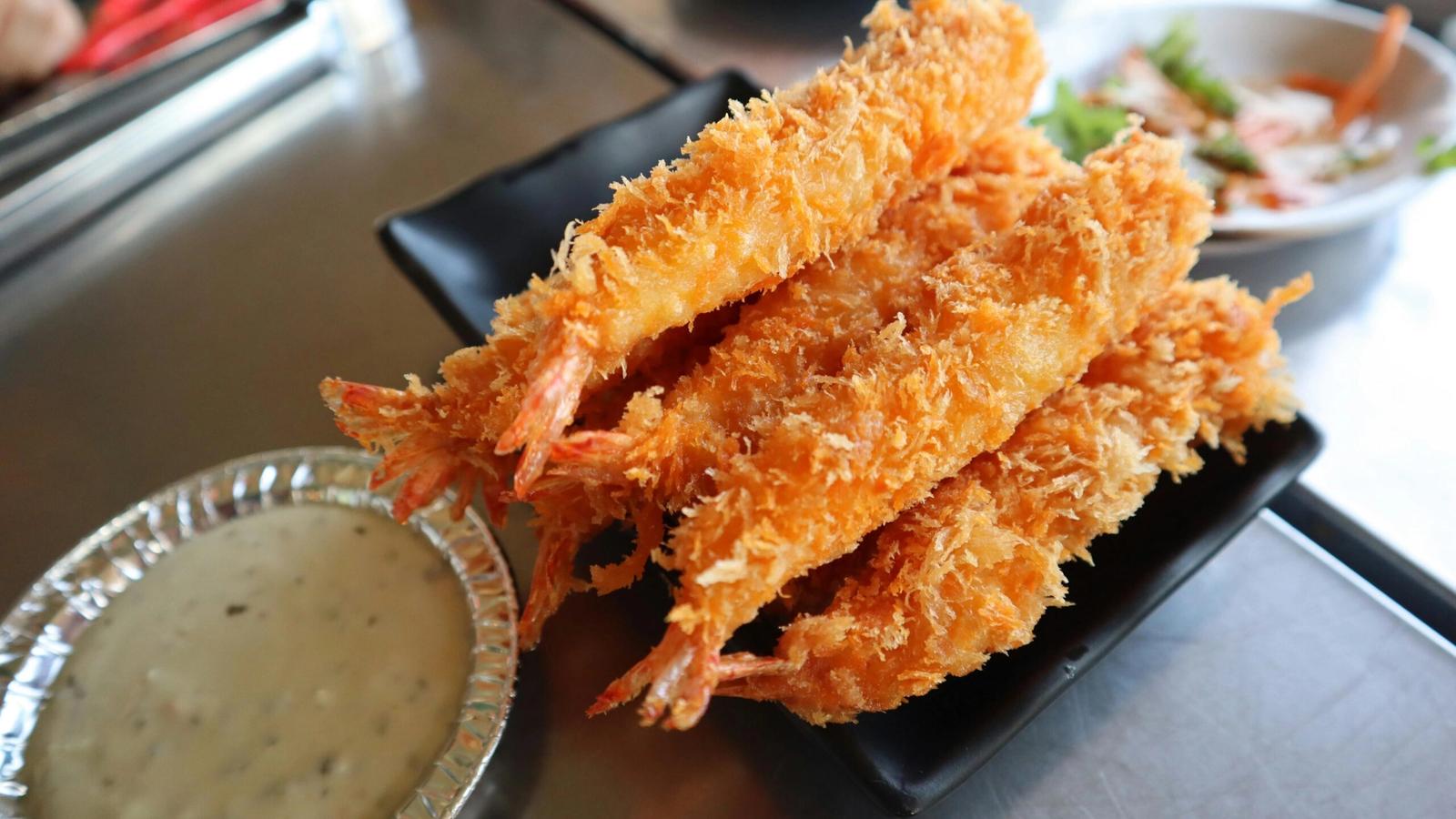 A black plate holds several pieces of golden, crispy fried shrimp from the Bubba Gump menu, with a foil dish of creamy dipping sauce nearby. In the background, there is a small plate of salad.