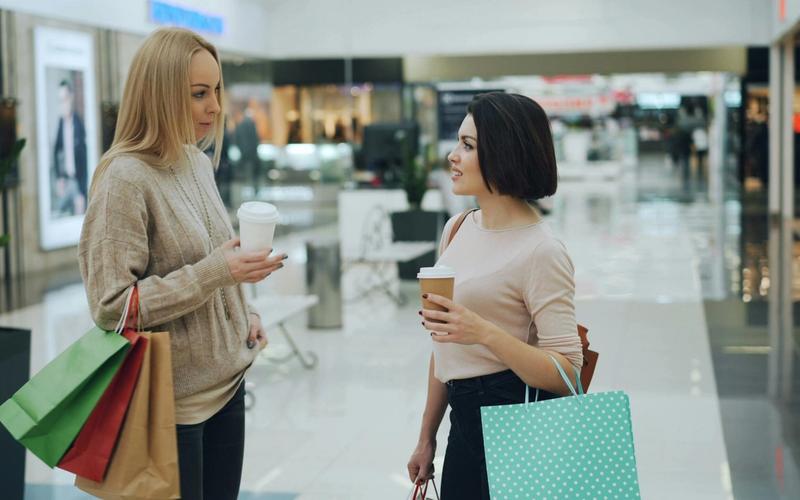 Two women holding shopping bags and coffee cups stand and talk in a brightly lit mall, surrounded by stylish shops in Gatlinburg. One woman has blonde hair and a beige sweater; the other has dark hair and a pale top. Storefronts and displays are visible behind them.