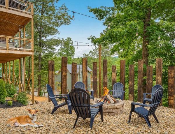 A cozy outdoor fire pit area with six black Adirondack chairs arranged in a circle on gravel, surrounded by trees and string lights, with a dog lying on the ground in the foreground.