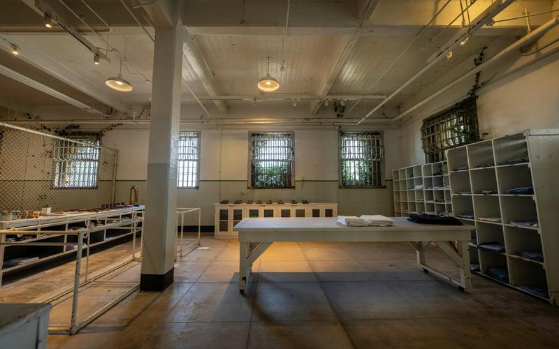 Large, empty room with barred windows, metal shelving, and tables with folded linens—like a prison laundry at Alcatraz East Crime Museum in Pigeon Forge. Industrial lighting and worn floors complete the scene. Tickets available for tours.