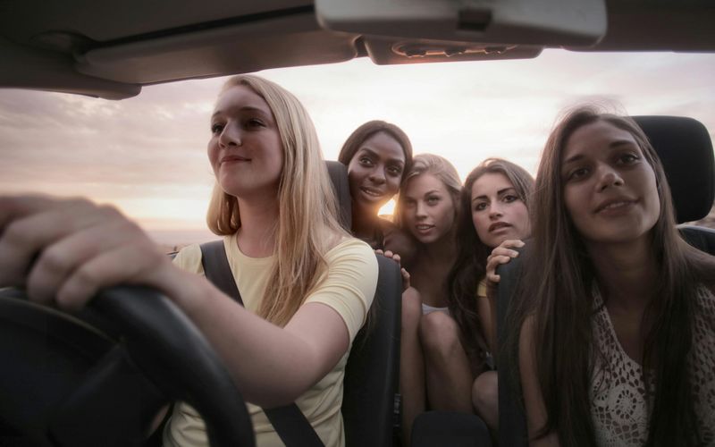 Five young women sit together in a car at sunset, with one driving and the others smiling and posing for a photo, capturing the cheerful spirit of a Girls’ Trip as they head toward one of the Best Places on their journey.