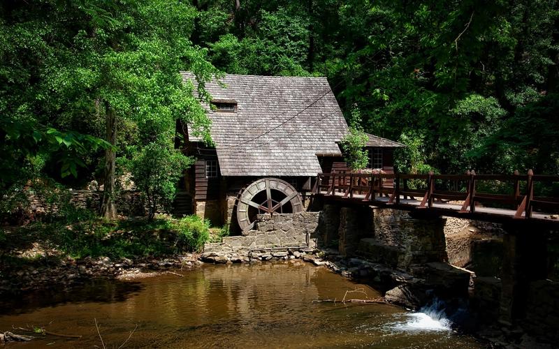 A rustic wooden watermill with a shingled roof sits by a stream, surrounded by dense green trees. Featured in many Old Mill Pigeon Forge guides, this scenic spot has a wooden bridge and sunlight filtering through the foliage.