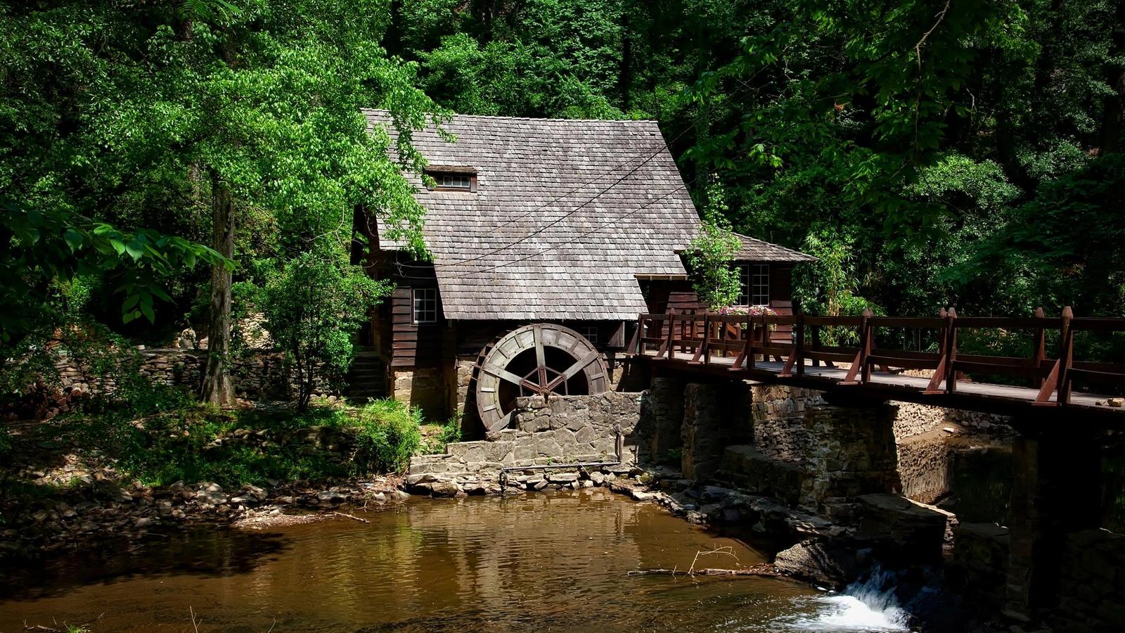 A rustic wooden watermill with a shingled roof sits by a stream, surrounded by dense green trees. Featured in many Old Mill Pigeon Forge guides, this scenic spot has a wooden bridge and sunlight filtering through the foliage.