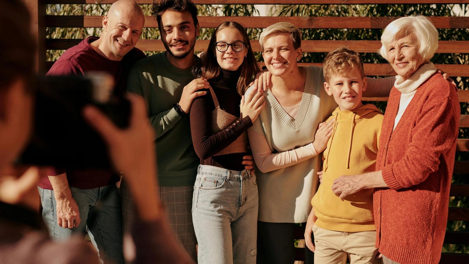 A group of six people of different ages stand close together, smiling for a family reunion photo outdoors. One person in the foreground is taking their picture. Trees and wooden slats are visible in the background, evoking the charm of the Smoky Mountains.