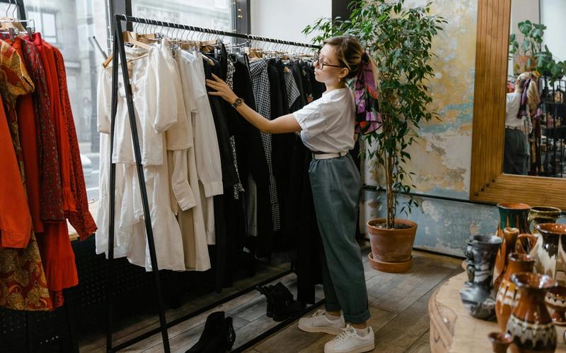 A woman wearing glasses, a white shirt, and jeans browses through clothes on a rack while shopping in Gatlinburg. The boutique features various garments, plants, and decorative items throughout the shop.